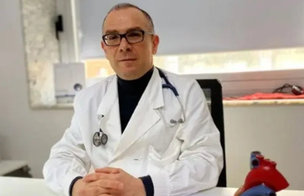 Davide Piano in camice bianco e stetoscopio, seduto a una scrivania bianca con un modello anatomico di cuore rosso e blu