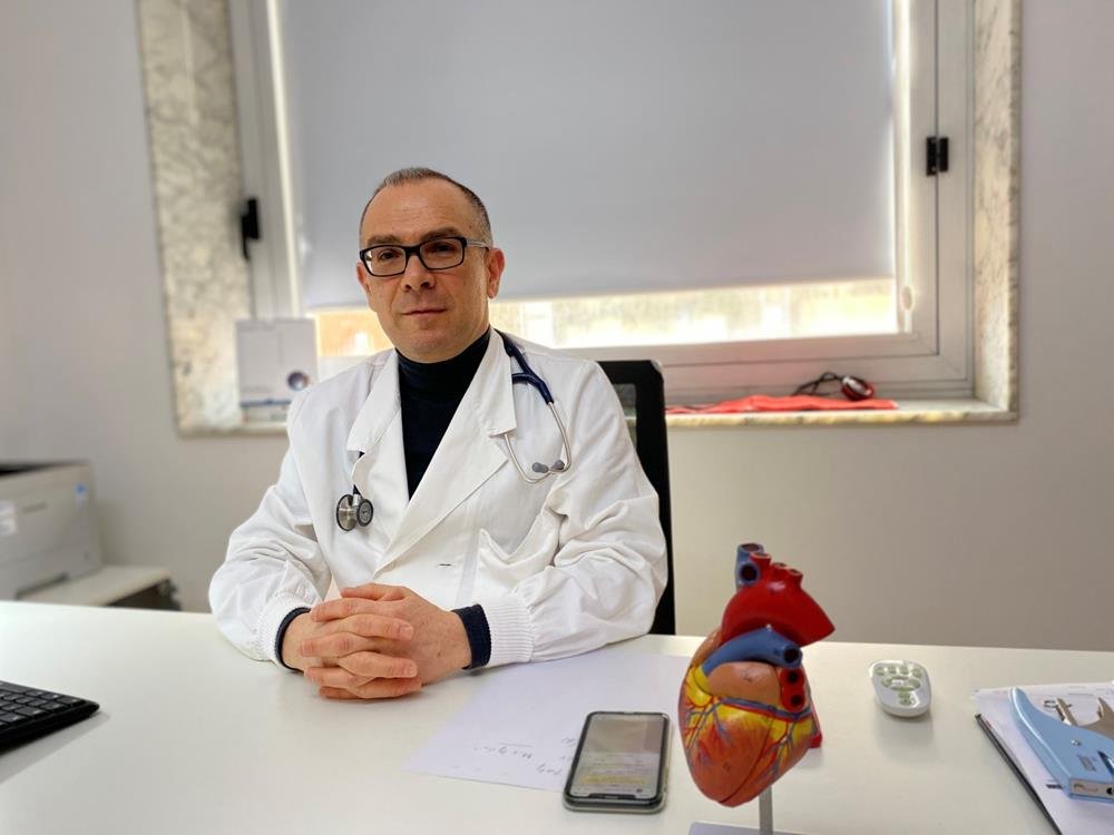 Medico con camice bianco e stetoscopio seduto alla scrivania, mani giunte, accanto a un modello anatomico del cuore