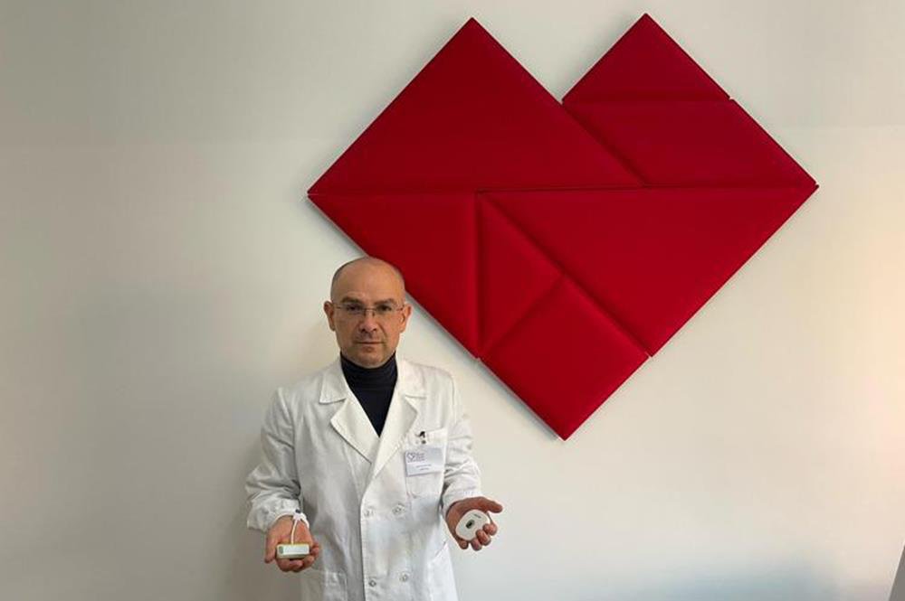 Medico in camice bianco mostra due piccoli registratori Holter davanti a un cuore di pannelli rossi sulla parete chiara